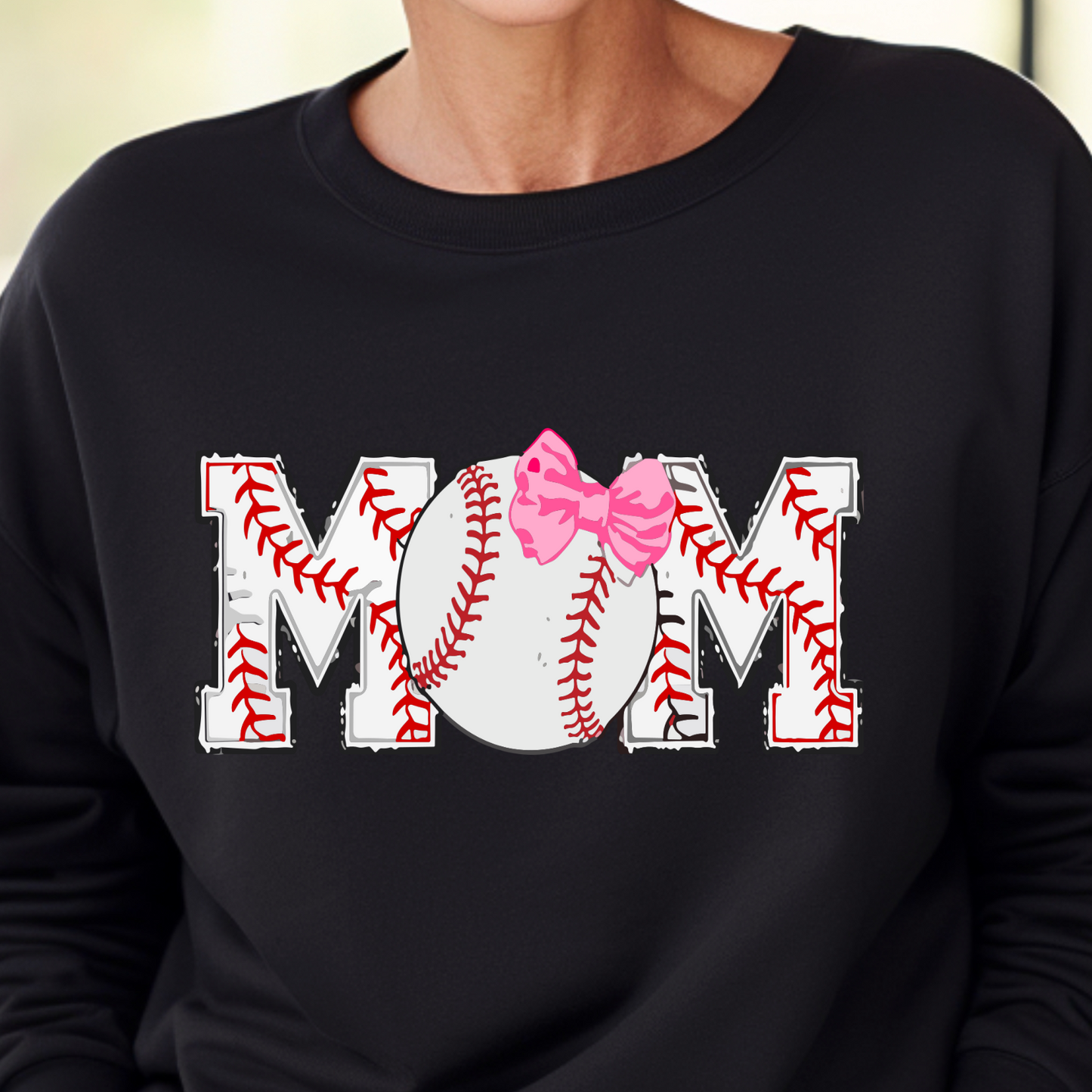 A woman wearing a black sweatshirt with the text 'MOM' and a pink bow design on it, smiling and standing in a house.