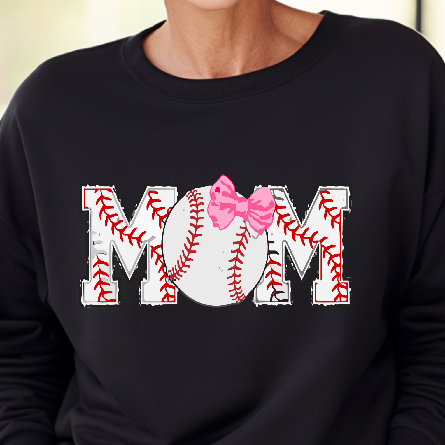 A woman wearing a black sweatshirt with the text 'MOM' and a pink bow design on it, smiling and standing in a house.