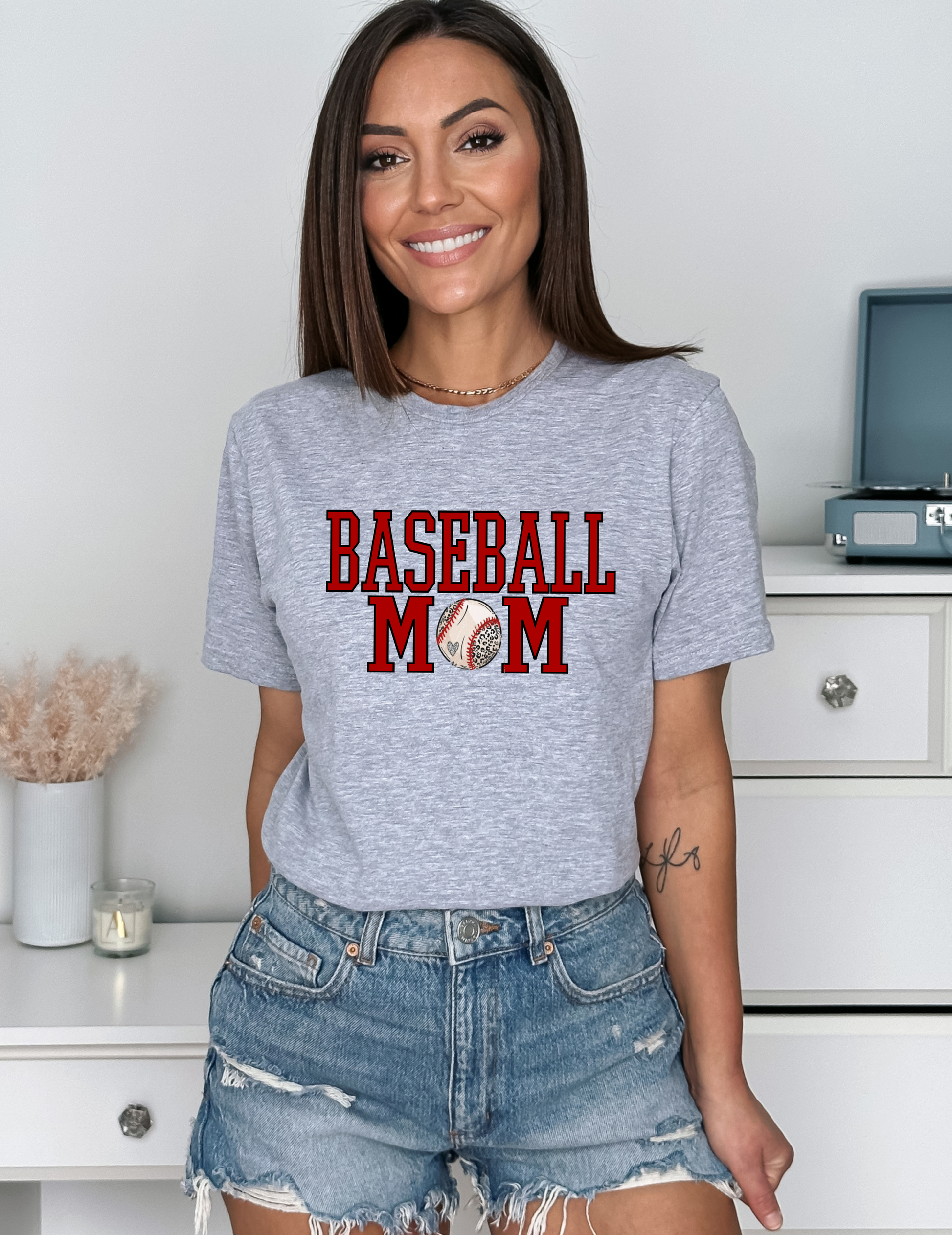 A woman wearing a gray t-shirt with 'BASEBALL MOM' written in red letters, paired with denim shorts.