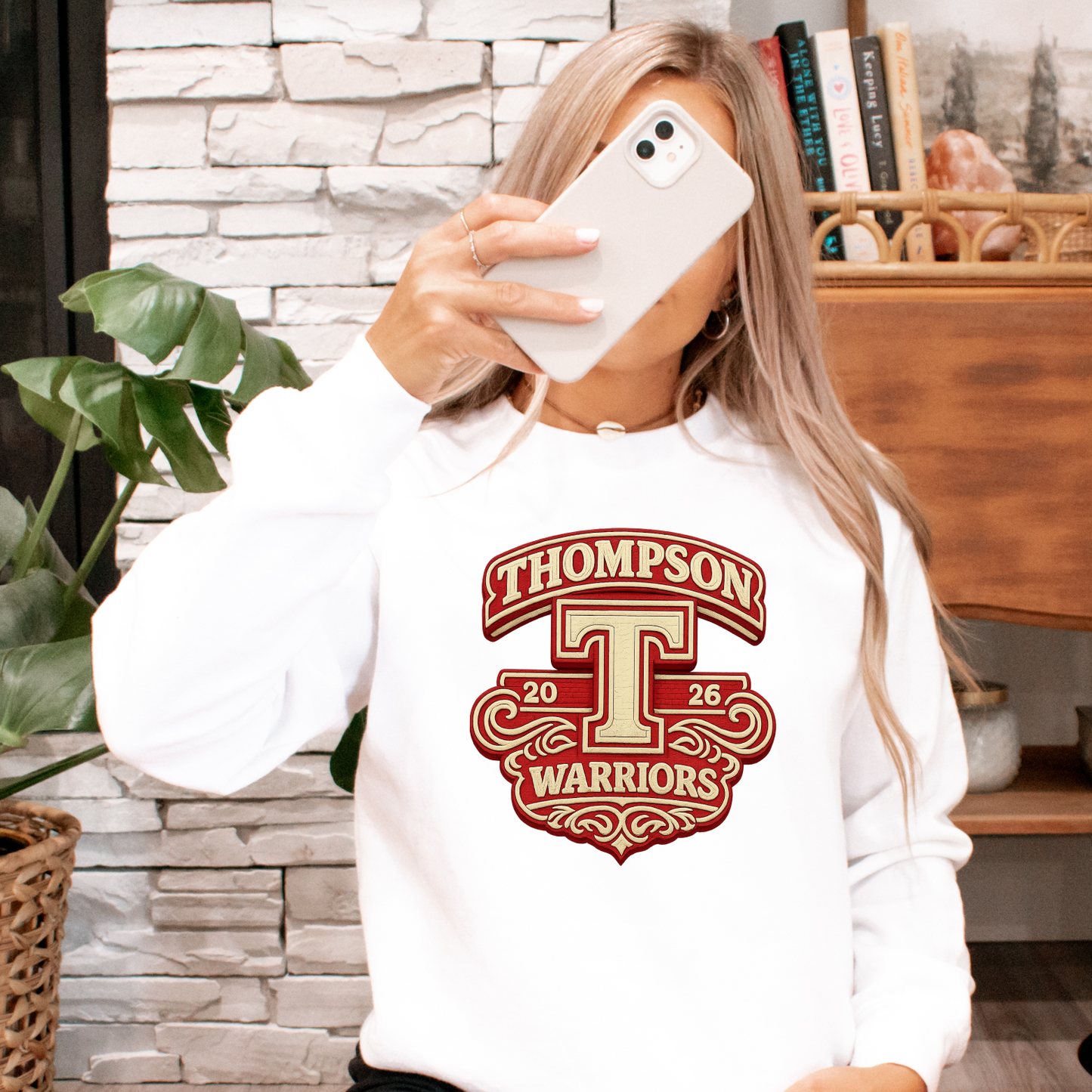 Person wearing a white sweatshirt with a school logo, sitting on the floor in a room with plants and a Victrola record player.